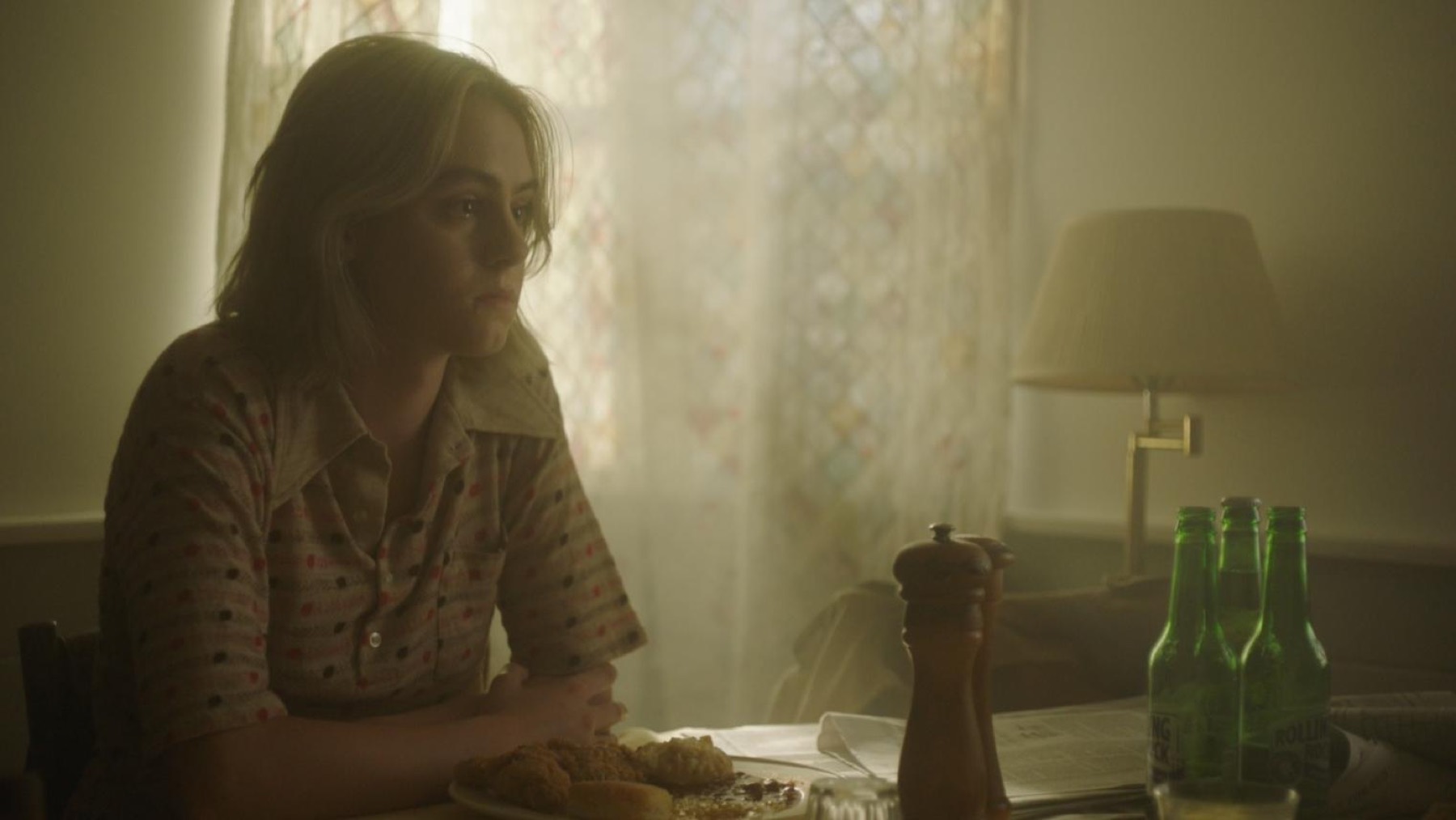 A young woman sits at a dining table in hazy afternoon light, a plate of fried food and green beer bottles in front of her.