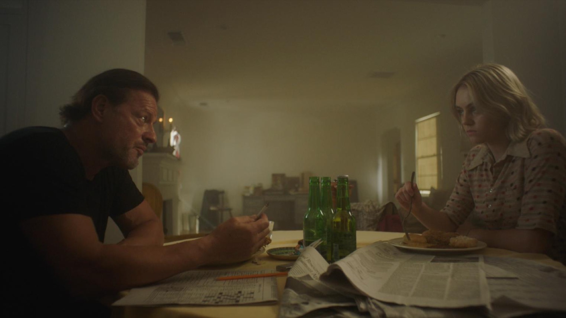 A man and a young woman sit at opposite ends of a dining table in dim light, beer bottles and a spread newspaper between them.