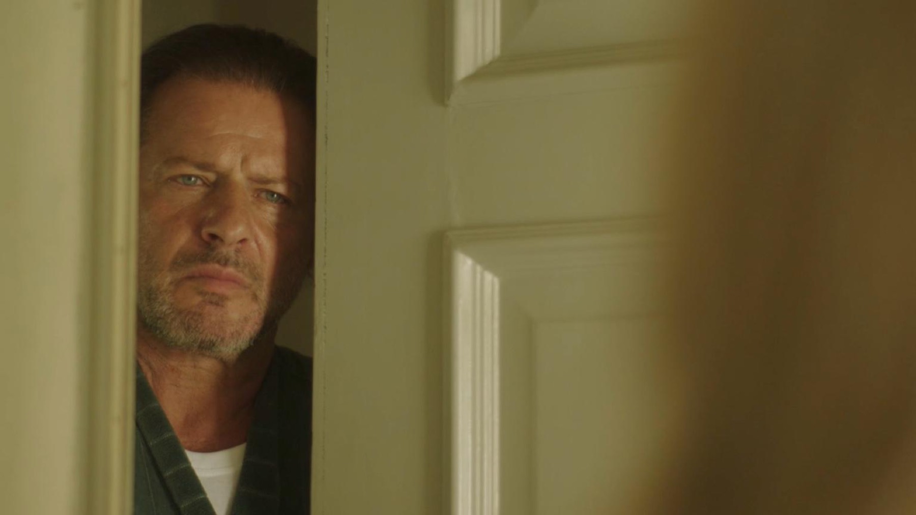 An older man framed in the narrow gap of a part-open door, watching.