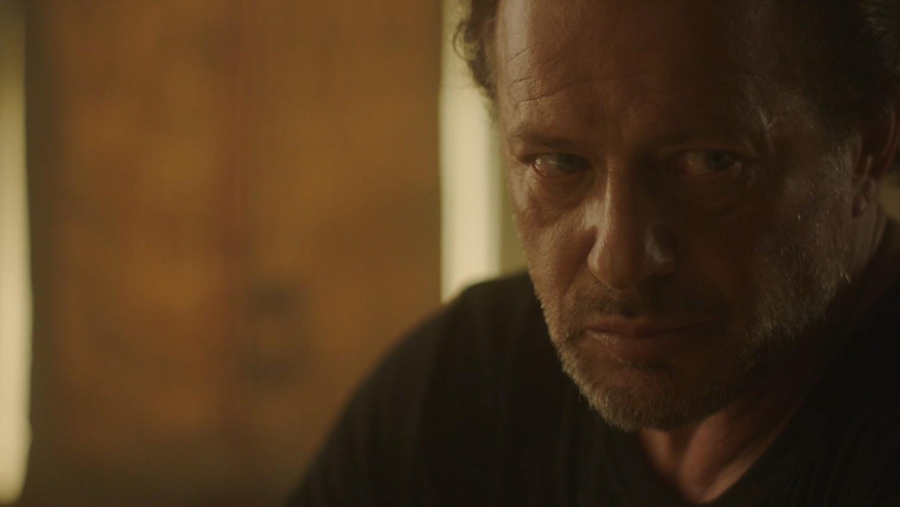 Close on an older man in a black t-shirt, half in shadow, looking off with a hard expression.