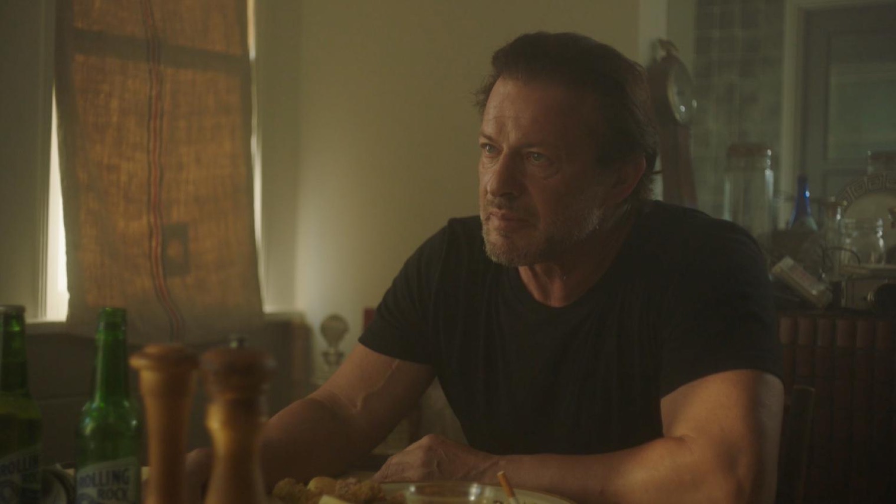 A man at a dining table in warm backlight, green beer bottles and pepper grinders in the foreground.