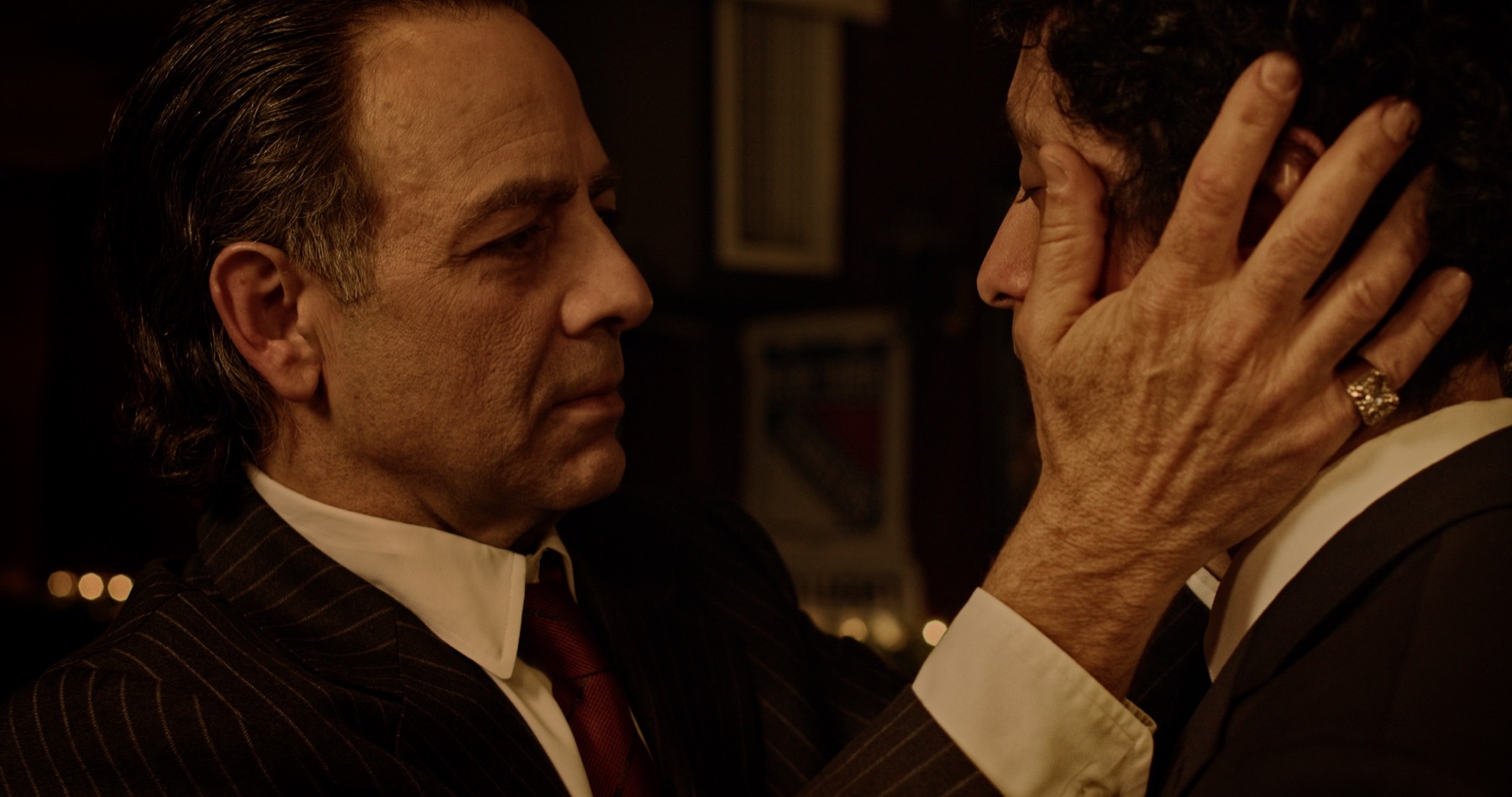 In a dim bar, an older man in a pinstripe suit cradles the face of a younger man in a dark suit.