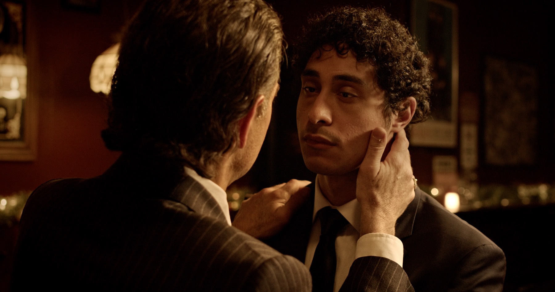 Over his shoulder, an older man in a pinstripe suit holds the younger man's face and straightens his collar in a dim bar.